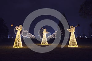 Three Christmas angels from luminous garlands illuminate the park walking area against the backdrop of round luminous