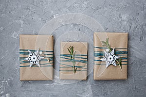 Three Christmas gifts in craft paper wrapped in blue thread, with white snowflakes and rosemary branches on a gray concrete