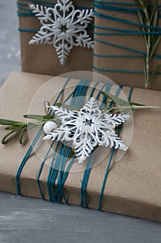 Three Christmas gifts in craft paper wrapped in blue thread, with white snowflakes and rosemary branches on a gray concrete