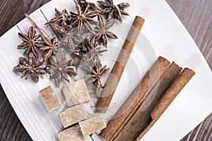 Three cinnamon sticks, anise stars and pieces of brown sugar lie on a white square saucer