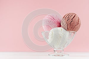 Three classic ice cream balls - strawberry, chocolate, creamy in elegant transparent glass bowl on white wood table, pastel pink.