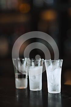 Three clean frosten short glasses with vodka stand on ice crumbs on bar stand.