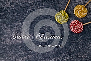 Three colored caramels on a stick in the upper corner on a dark blue metallic background with christmas greeting. Top view