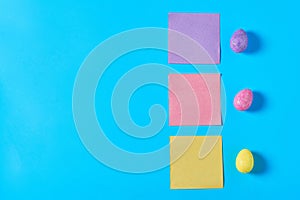 Three colorful eggs with glitters near blank square paper stickers lies on blue desk on kitchen