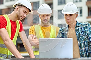 Three concentrated men in protective helmets standing before a laptop