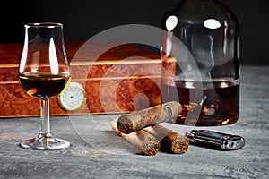 Three Cuban cigars on a stone table with a lighter and a wooden humidor with a glass and a bottle of Whiskey photo