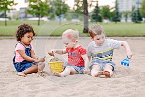 Three cute Caucasian and hispanic latin toddlers babies children sitting in sandbox playing with plastic colorful toys