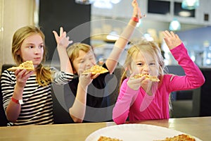 Three cute funny siblings eating slices of pizza at indoor restaurant or cafe