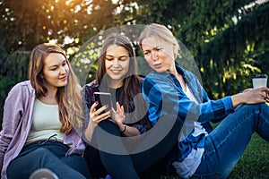 Three cute girls relax and socialize on the lawn in summer park. Young women sit on the green grass among the trees and look