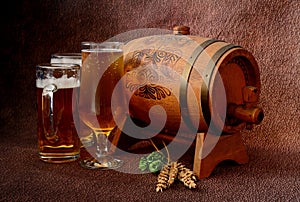 Three different glasses with light beer, next to a wooden barrel, hops and barley ears on a brown abstract background