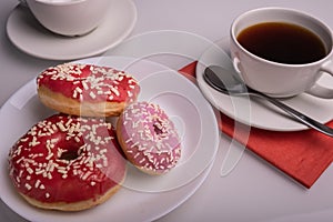 Three donuts coated with pink icing with sprinkles and cup of coffee on the table