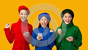 Three Excited Girls Holding Smartphones Gesturing Yes Over Yellow Background