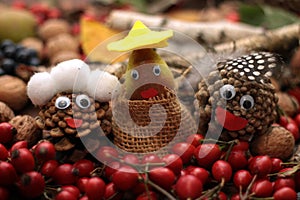 Three faces made of cones with eyes and mouth, head cover surounded by falll items as rose hip, nuts, grapes, leaves