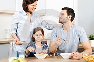 Three family members have delicious healthy breakfast at kitchen, eat cornflakes with milk, enjoy togetherness and