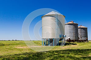 Three farm grain silos for agriculture. New and old silos. Placed next to the cultivated field. Countryside in south america.