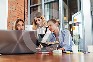 Three female college students doing homework together using one laptop and lecture notes sitting at desk in study room