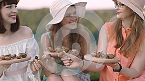 Three female friends, sitting on the blanket in meadow and holding wooden plates with escargots