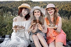 Three female friends, sitting on the blanket in meadow and holding wooden plates with escargots