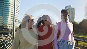 Three female friends walking and talking arm in arm outdoors in the city during a sunny day.