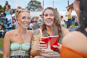 Three Female Friends Wearing Glitter Having Fun At Summer Music Festival Doing Cheers With Drinks