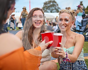 Three Female Friends Wearing Glitter Having Fun At Summer Music Festival Doing Cheers With Drinks