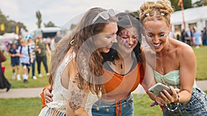 Three Female Friends Wearing Glitter Looking At Mobile Phone At Summer Music Festival 