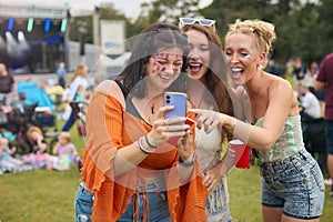 Three Female Friends Wearing Glitter Looking At Mobile Phone At Summer Music Festival Holding Drinks