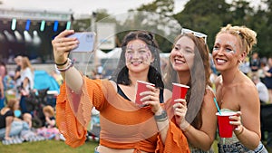 Three Female Friends Wearing Glitter Posing For Selfie At Summer Music Festival Holding Drinks