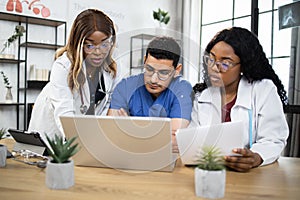 Three multiracial doctors having conference at hospital, working with laptop pc