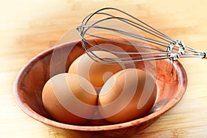 Three fresh brown eggs in a bamboo bowl and a wire whisk on a wooden table. Concept of baking/cooking