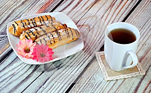 Three fresh eclairs decorated with flower buds and chocolate on a plate stand next to a white high mug of tea