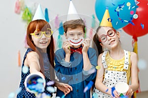 Three friendly children in festive cone caps and big eyewear, stand in decorative room with balloons, have fun together