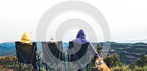 three friends sit in camping chairs on top of a mountain, travelers enjoy nature and cuddle with dog, tourists look into distance