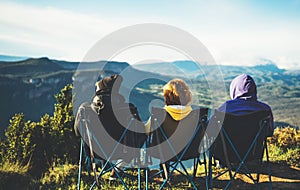 Three friends sit in camping chairs on top of a mountain, travelers enjoy nature and cuddle, tourists look into distance on back