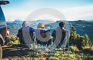 Three friends sit in camping chairs on top of a mountain, travelers enjoy nature and cuddle, tourists look into distance on back