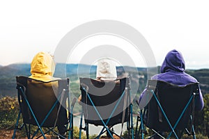 Three friends sit in camping chairs on top of a mountain, travelers enjoy nature and cuddle, tourists look into distance on back