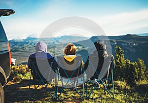 Three friends sit in camping chairs on top of sun mountain, travelers enjoy nature, tourists look into distance on background
