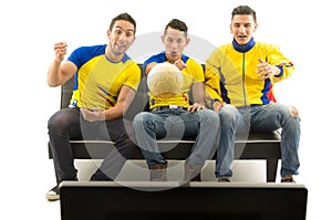 Three friends sitting on sofa wearing yellow sports shirts watching television with enthusiasm, golden ball flying in