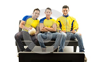 Three friends sitting on sofa wearing yellow sports shirts watching television with enthusiasm, white background, shot