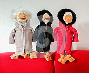 Three geese stuffed animals with weatherproof jackets sit on a red sofa in front of a white wall