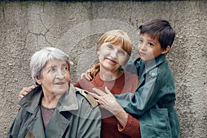 three generations grandmother great grandmother grandson are standing on street against a gray wall