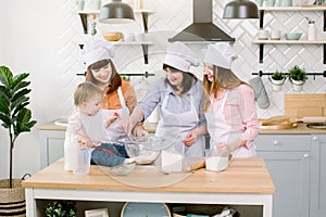 Three generations of women in white aprons are playing and laughing while kneading the dough in the kitchen, mix flour