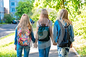 Three girl schoolgirl girlfriends. They hold each other`s hands. Summer in city. Back view. They walk down the street to