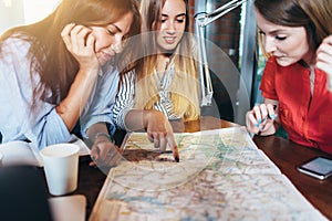 Three girlfriends planning their vacation sitting at table around map choosing the destination