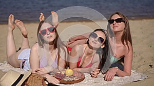 Three girlfriends in swimsuits and sunglasses are posing, lying down on a coverlid on a sandy beach by the river. The