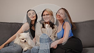 Three girlfriends are taking photos, sitting on the sofa. The girl sets the self-timer on the camera and sits down with