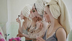 Three girlfriends with towels on their heads are doing morning makeup while standing in front of the mirror in the