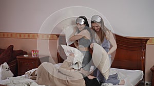 Three girlfriends wearing sleep masks on their heads had a comic pillow fight while kneeling on a bed in the bedroom