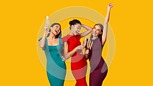 Three Girls Celebrating Drinking Sparkling Wine And Dancing, Yellow Background