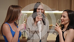 Three girls clink glasses of white sparkling wine, smile and drink while standing in the kitchen in a modern apartment.